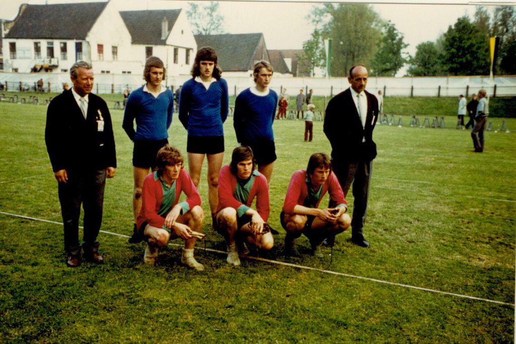 1972-finale-jong-nederland-sint-jabik-wommels-scaled - kaatsnieuws.com Vintage soccer team photo: six players posing on a grassy field with two suited men, three standing in blue shirts and three kneeling in red shirts, houses in the background.