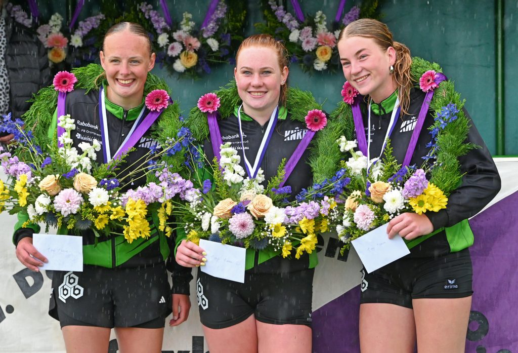 1e Prijs Winnaarsronde Dames 1ste Klas - kaatsnieuws.com Three athletes on a podium wearing green and black, each with a flower wreath and medal ribbons, holding notes.