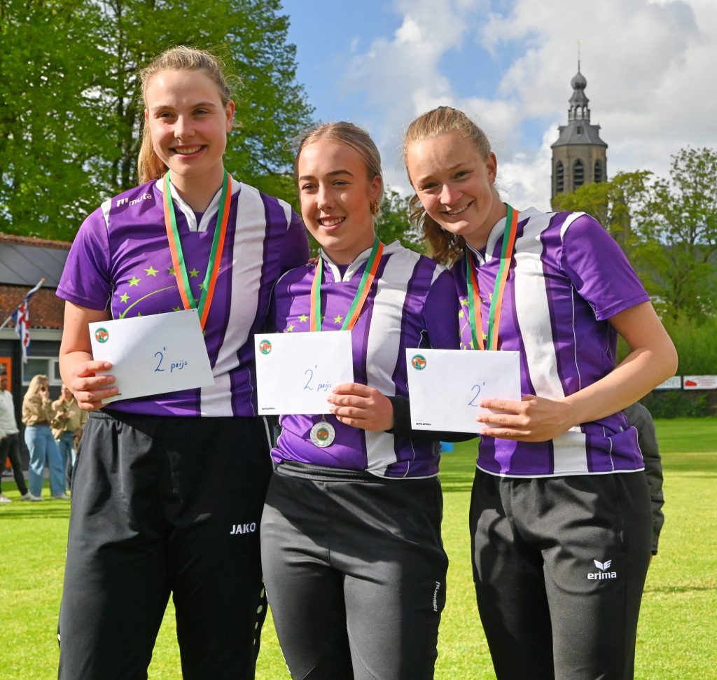 Three female athletes in purple and white uniforms with medals and prize certificates, posing outdoors on a sunny day.