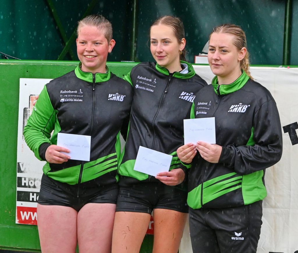 2de Prijs Herkansing Dames 1ste Klas - kaatsnieuws.com Three female athletes in green and black uniforms pose with envelopes on a podium after a race event.