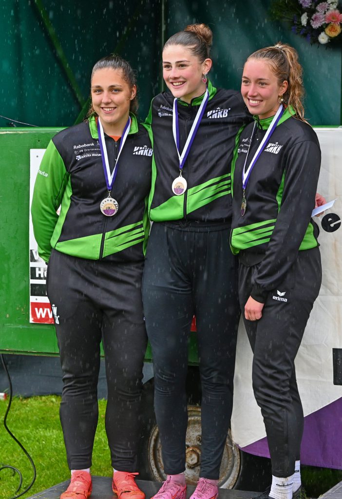 2de Prijs Winnaarsronde Dames 1ste Klas-2 - kaatsnieuws.com Three female athletes on a podium wearing matching green and black outfits, each with medals, smiling after the event.