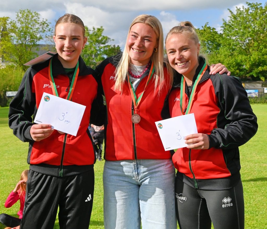 Three smiling women in red-and-black jackets pose together outdoors, each wearing a medal on a green field.