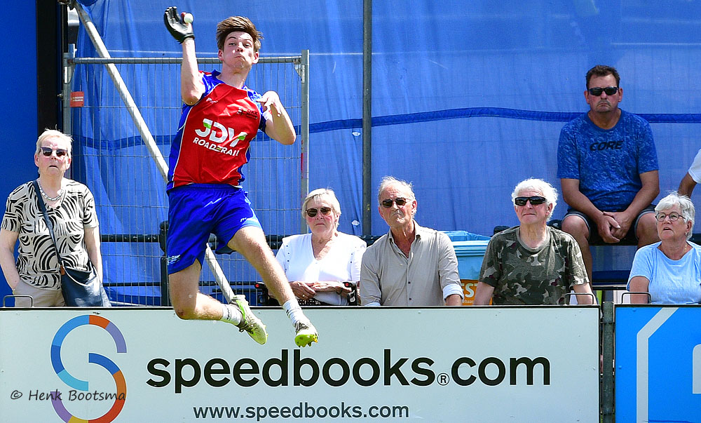 Male long jumper in red and blue uniform mid-air over a barrier with spectators watching behind a fence and a speedbooks.com banner visible.