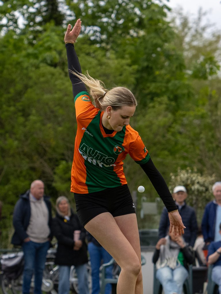 Female athlete in an orange-green jersey raises her arm during an outdoor event, with spectators watching in the background.
