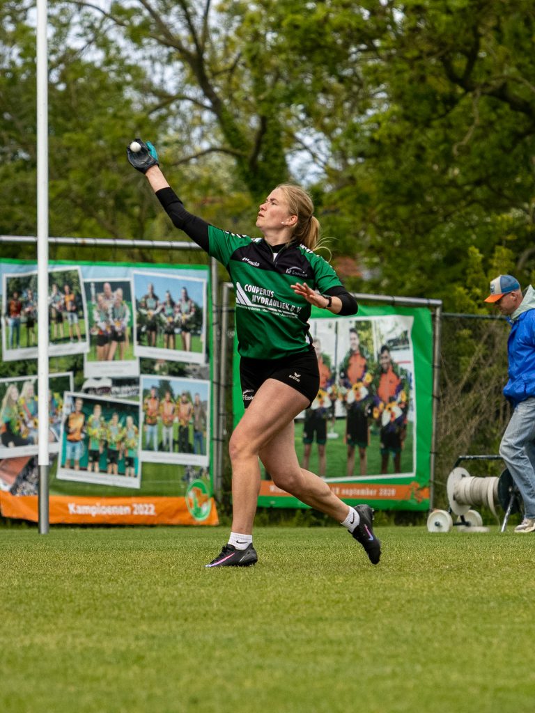 Female athlete in green and black uniform throws a ball on a grassy field, with banners and spectators in the background.