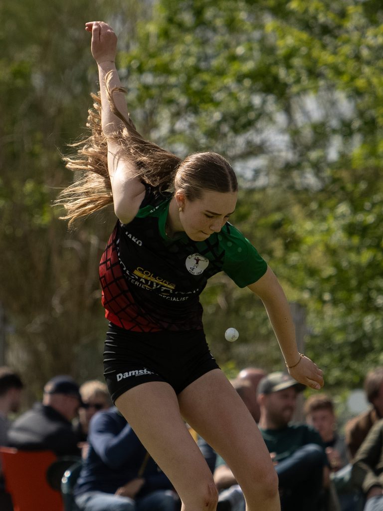 Female athlete in a green and black uniform mid-throw, arm raised and a ball in the air, with spectators watching in the background.