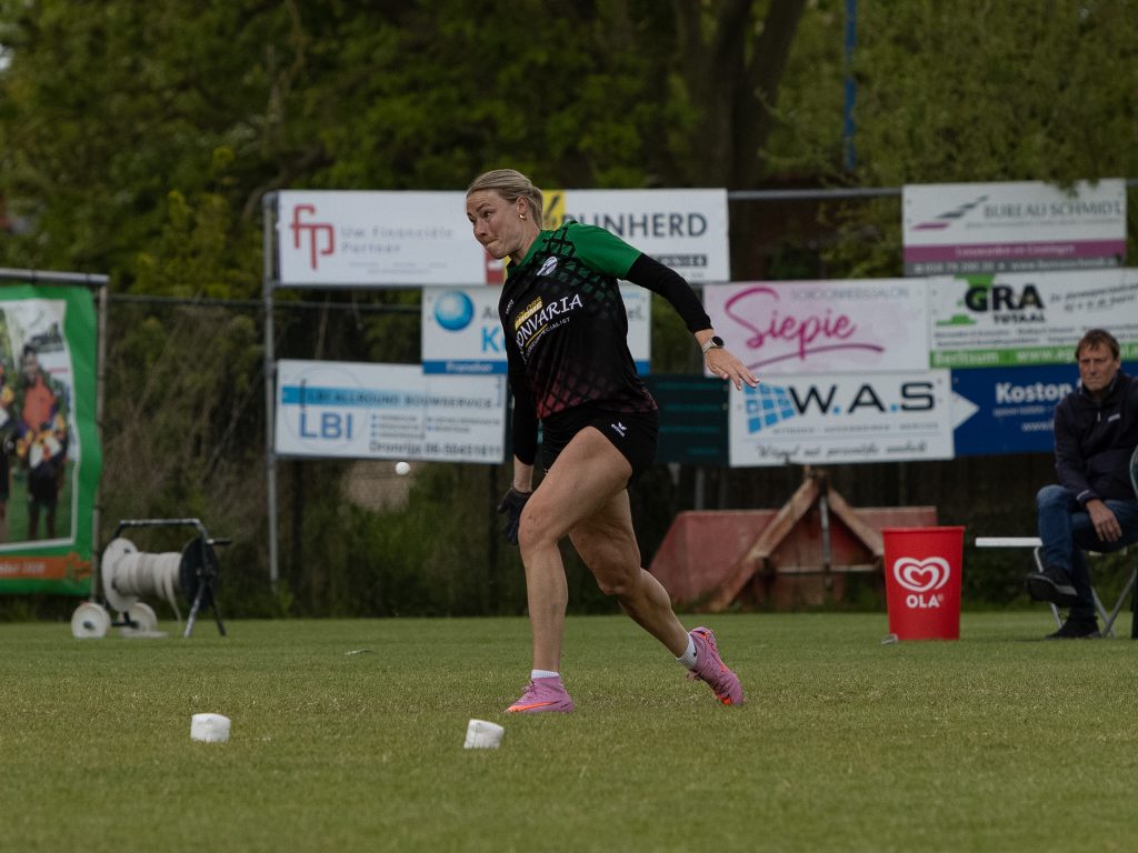 Female athlete in a green jersey sprinting on a grassy field, sponsor boards in the background during a race.