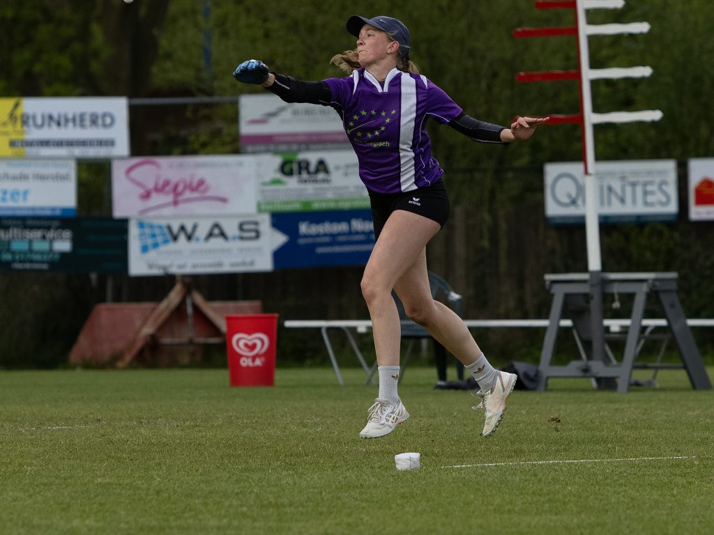 Female athlete in purple jersey throws a discus/shot put on a sports field, mid-throw with audience boards in the background.