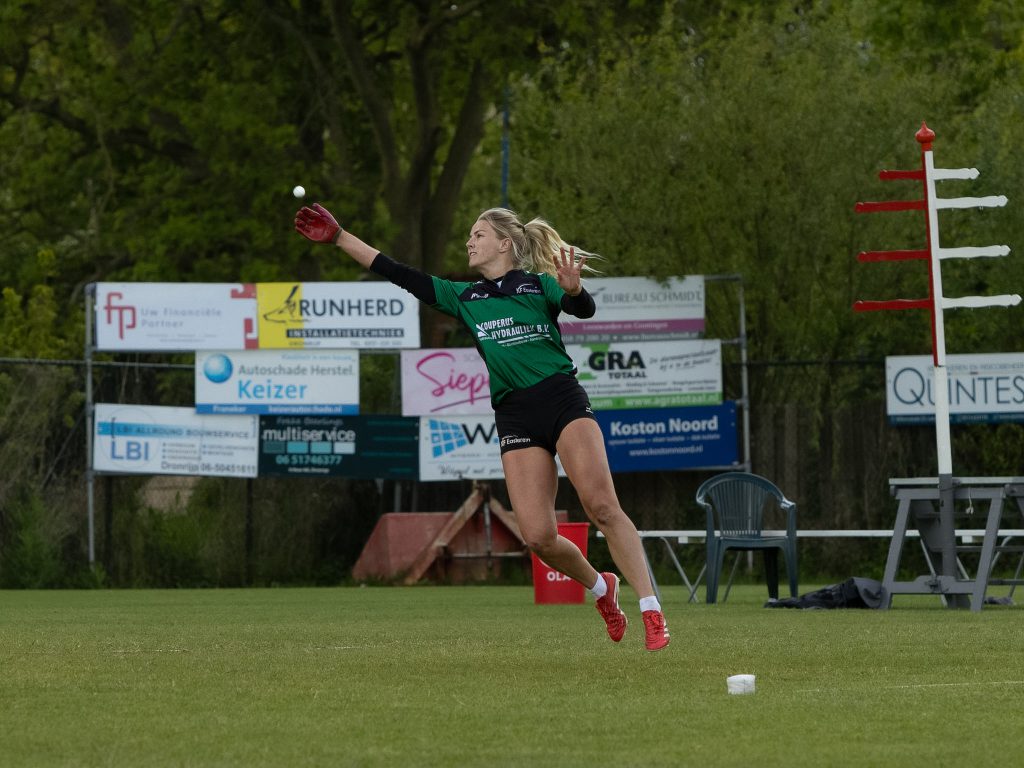 Female athlete in a green jersey throws a white ball on a grassy field with advertising boards in the background.