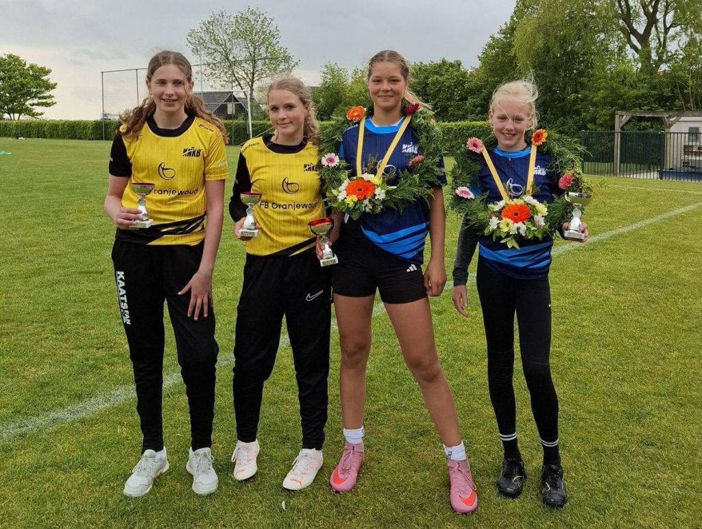 A_pup-easterein - kaatsnieuws.com Four young girls on a grassy field pose with trophies after a competition; two wear yellow jerseys and two wear blue outfits with flower garlands around their necks.