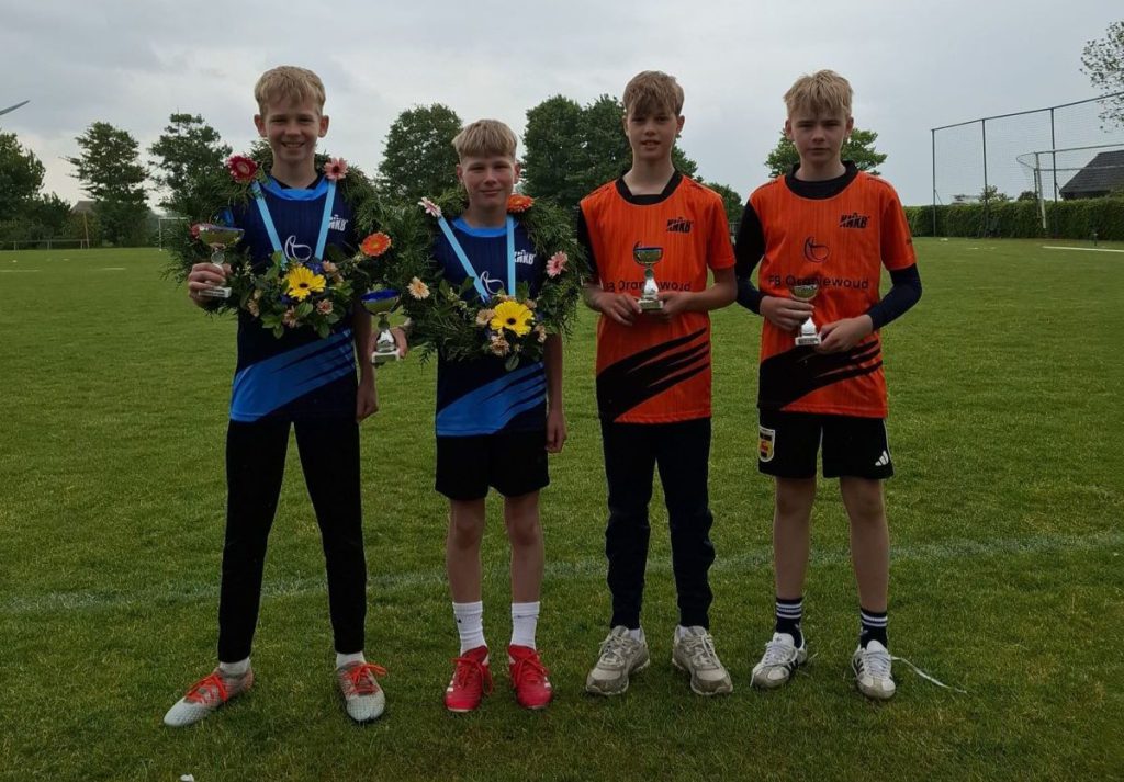 Apupjong - kaatsnieuws.com Four boys pose on a grassy field after a sports event; two in blue uniforms hold floral wreaths and trophies, two in orange shirts hold smaller trophies.