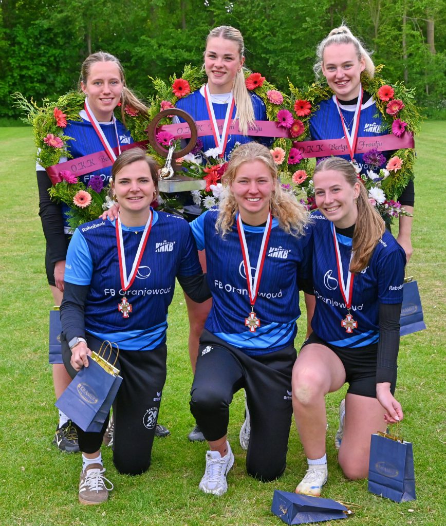 Six athletes in blue uniforms pose on a grassy field with flower wreaths, medals, and trophies to celebrate a win.