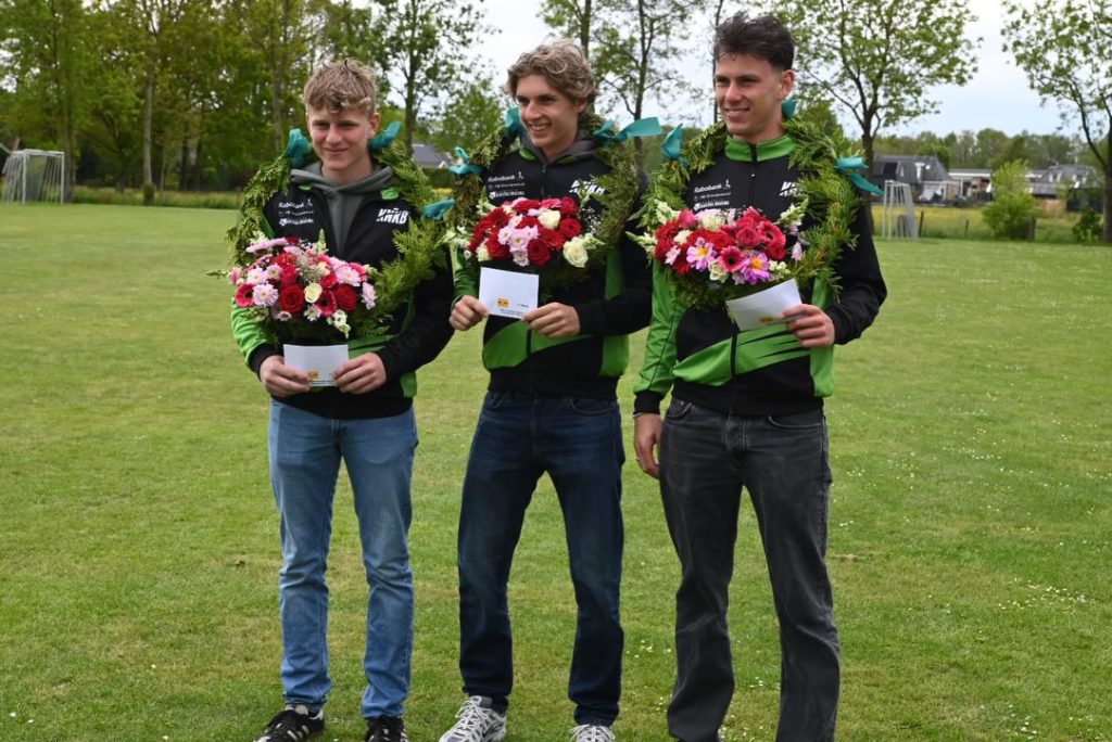Broeksterwald 1 - kaatsnieuws.com Three young men in black-green jackets wear floral wreaths, holding bouquets and cards on a grassy field after a ceremony.