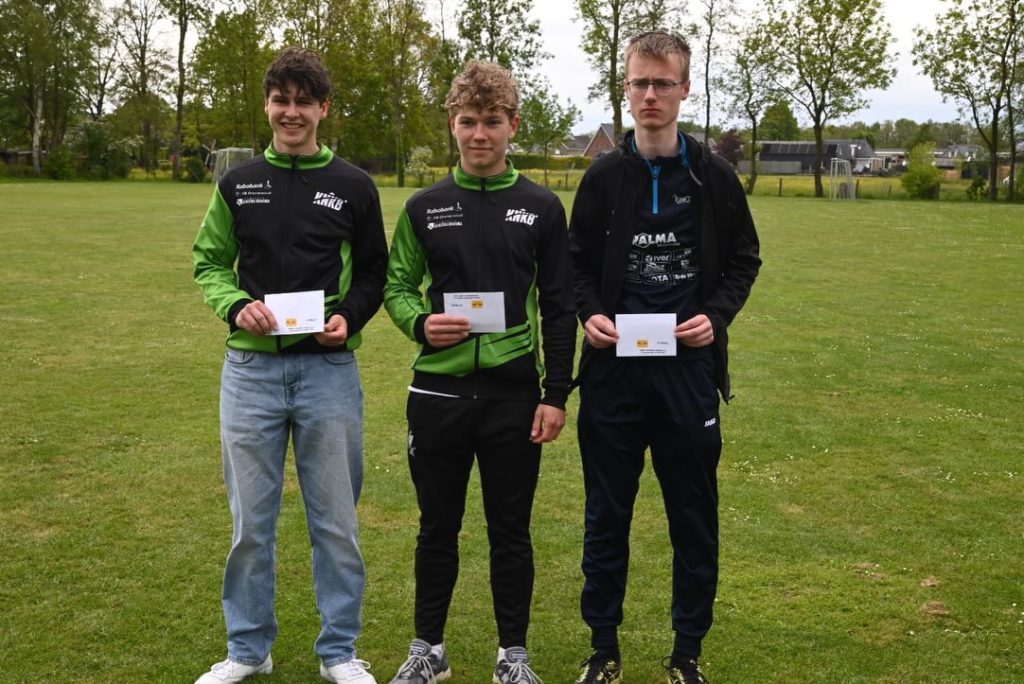 Broeksterwald herkansing - kaatsnieuws.com Three young men stand on a grassy field, each holding white envelopes and looking at the camera; two wear green-and-black jackets, one wears a black jacket.