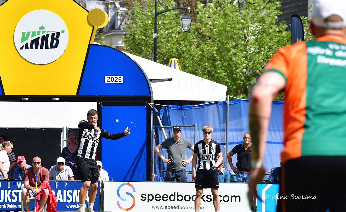 Soccer players in black-and-white striped jerseys compete outdoors, a man kicks the ball while spectators watch. Advertising banners line the area.