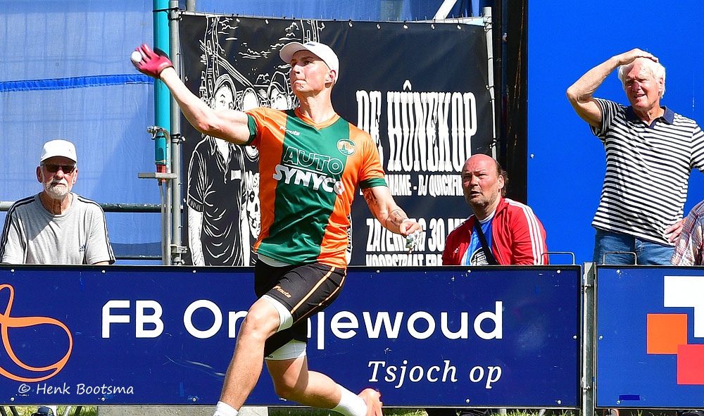 Athlete in green and orange jersey mid-throw on a sunny sports field, with spectators behind advertising boards.