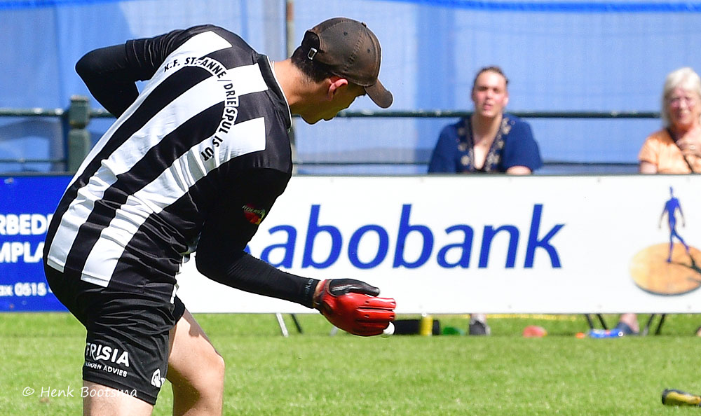 Soccer goalkeeper in black-and-white striped kit bends forward, wearing red gloves, on a grassy field with spectators and a Rabobank ad backdrop.