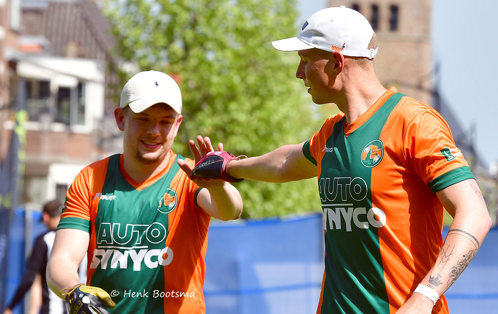 Two baseball or cricket players in orange-and-green uniforms high-fiving during practice or game outdoors.