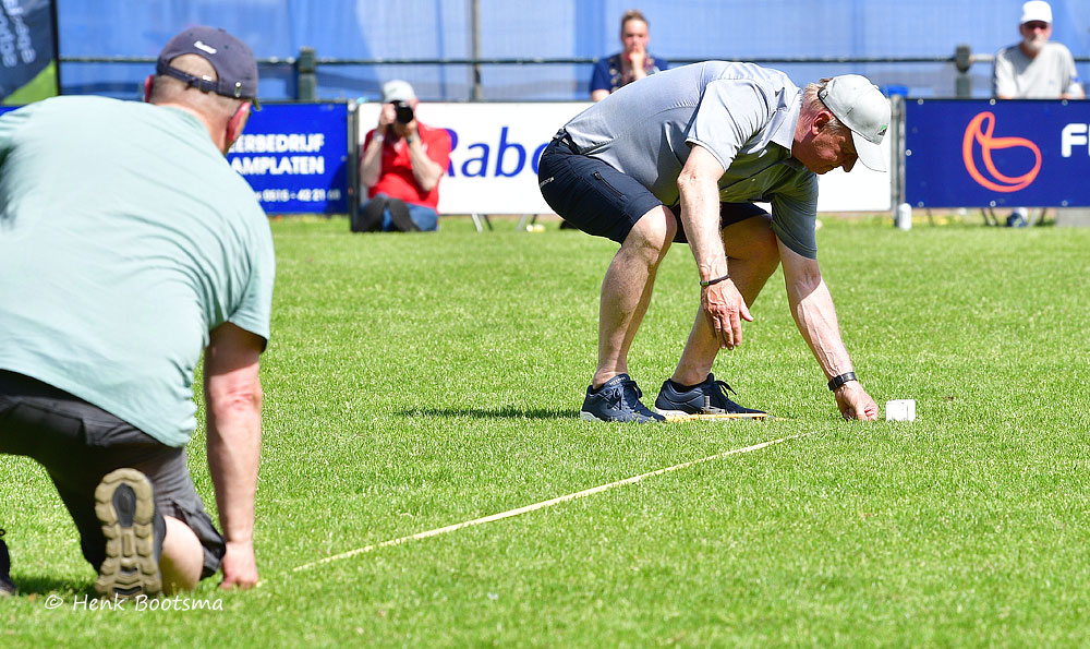 Two men on a grassy field set up a line for a lawn bowls shot, with a white jack placed on the ground and spectators in the background