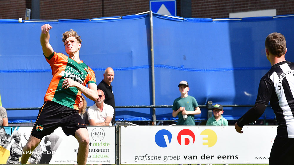 Young male player in an orange-and-green jersey throws a ball in an outdoor court while an opponent in a black-and-white kit watches, with spectators behind a blue barrier.