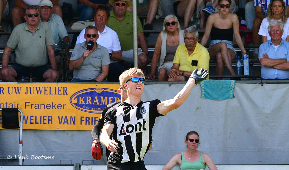 Soccer player in black-and-white stripes raising his arm while running in a stadium with spectators behind and a yellow sponsor banner.