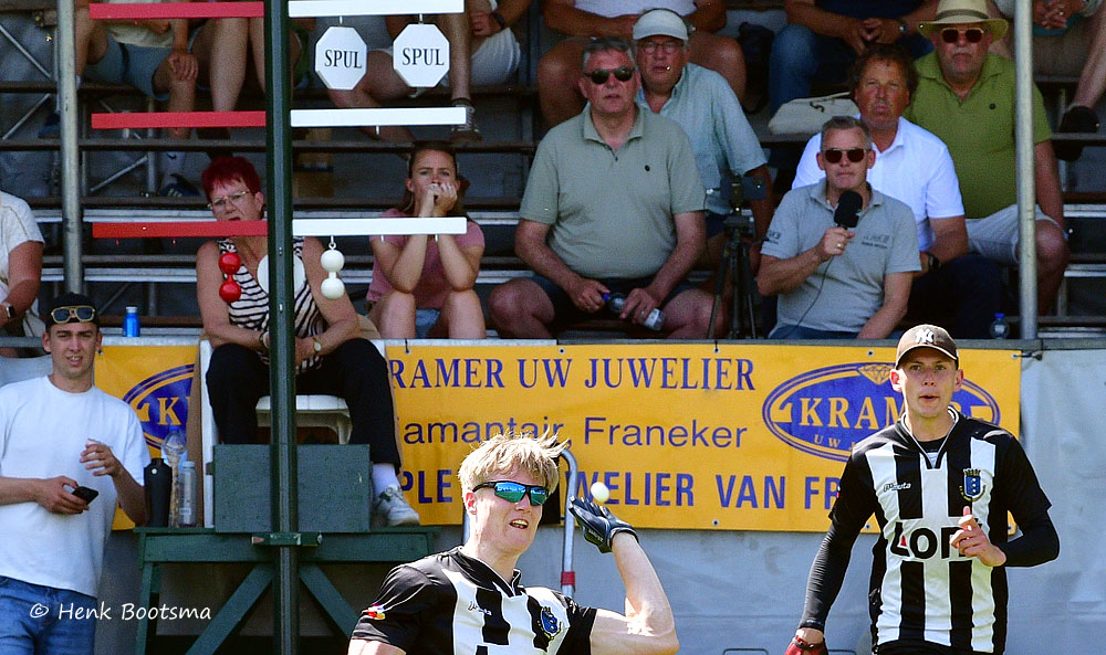 Two men in black-and-white striped jerseys playing a ball game, with spectators behind a yellow banner reading 'KRAMER UW JUWELIER'.