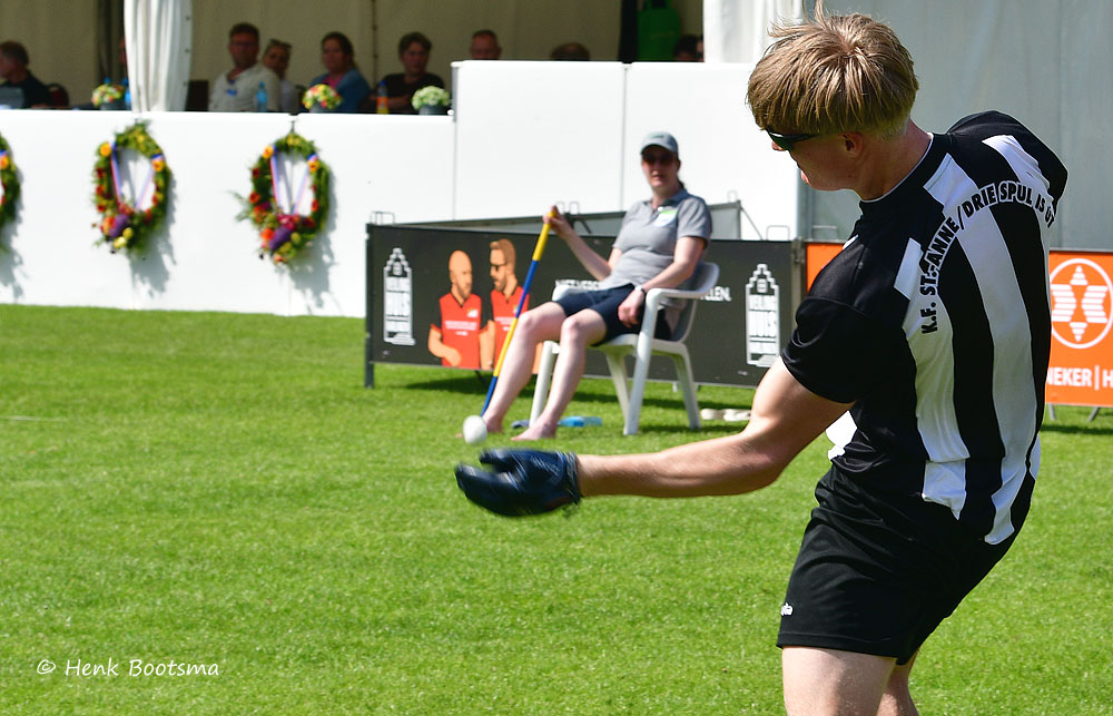 Lawn bowls player in a black-and-white striped jersey delivering a ball on a green field, with spectators and banners in the background.