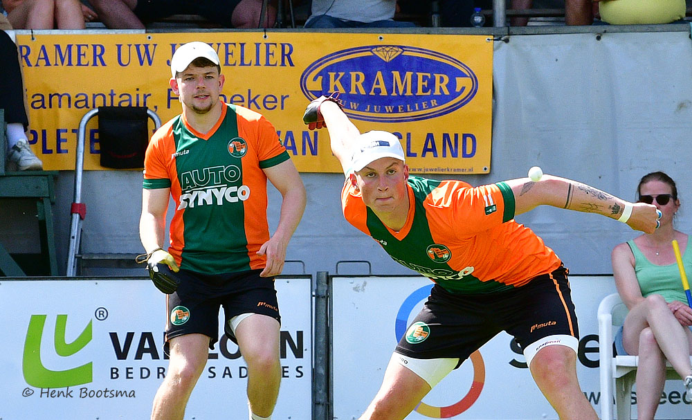 Two field hockey players in orange and green uniforms chase a ball during a match, with a sponsor banner in the background.