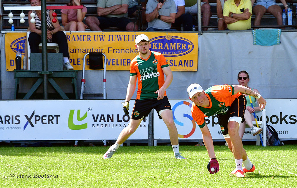 Two field hockey players in orange and green uniforms sprint on a sunny grass field, with spectators and banners in the stands behind them.] ,