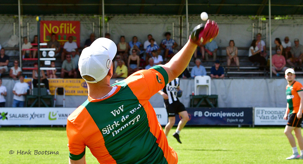 Handball player in orange and green jersey throwing a ball mid-air during a match, with spectators seated in the stands behind.