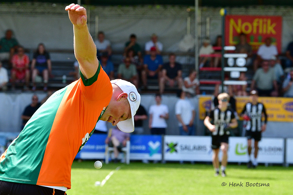Athlete in an orange and green shirt throws a small ball during an outdoor game, with spectators watching from the stands in the background.