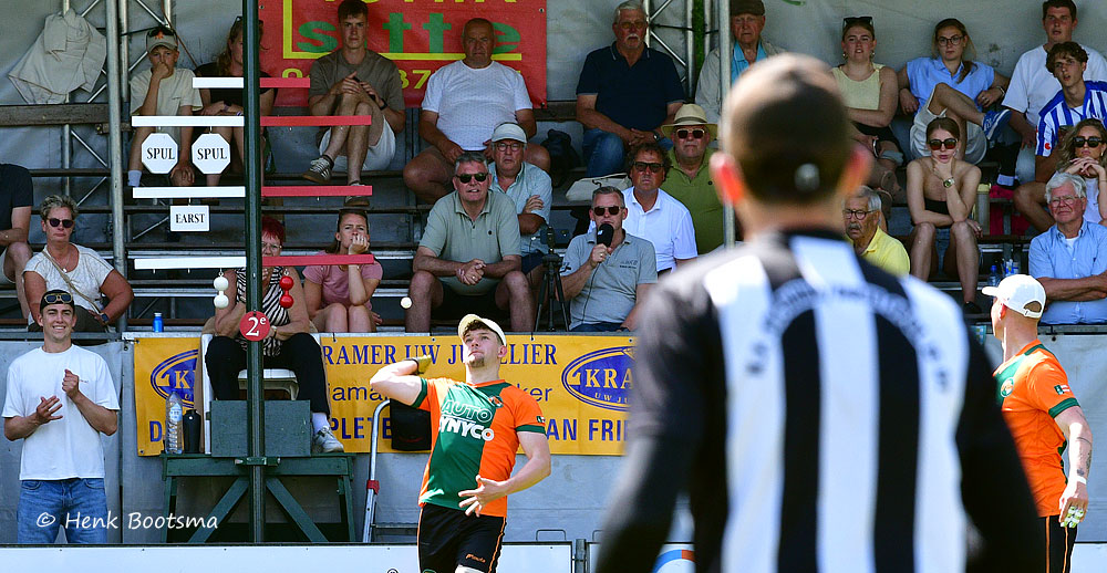 Outdoor sports match: player in orange and green uniform raises an arm near the net as a referee in a black-and-white striped shirt stands in the foreground, with a crowd watching from stands behind a yellow banner.