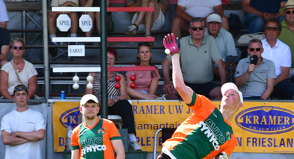 Two players in orange and green uniforms play a throwing sport on an outdoor court as one player releases a ball mid-air; spectators watch from bleachers behind a yellow banner.