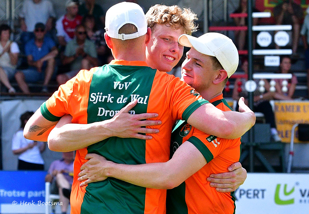 Two teammates in orange and green jerseys embrace in celebration on a sports field with spectators in the stands behind them.
