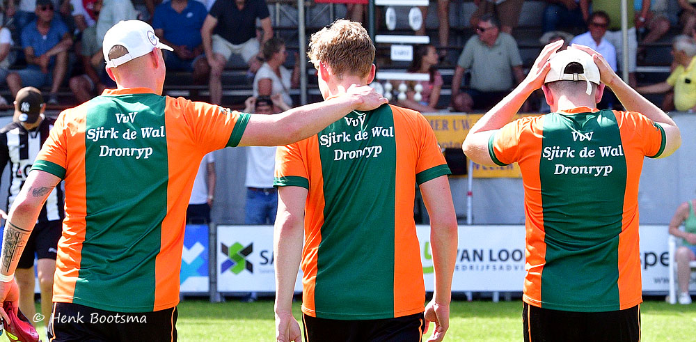 Three players in orange and green jerseys stand with arms around each other on a grass field, facing away from the camera as spectators watch.