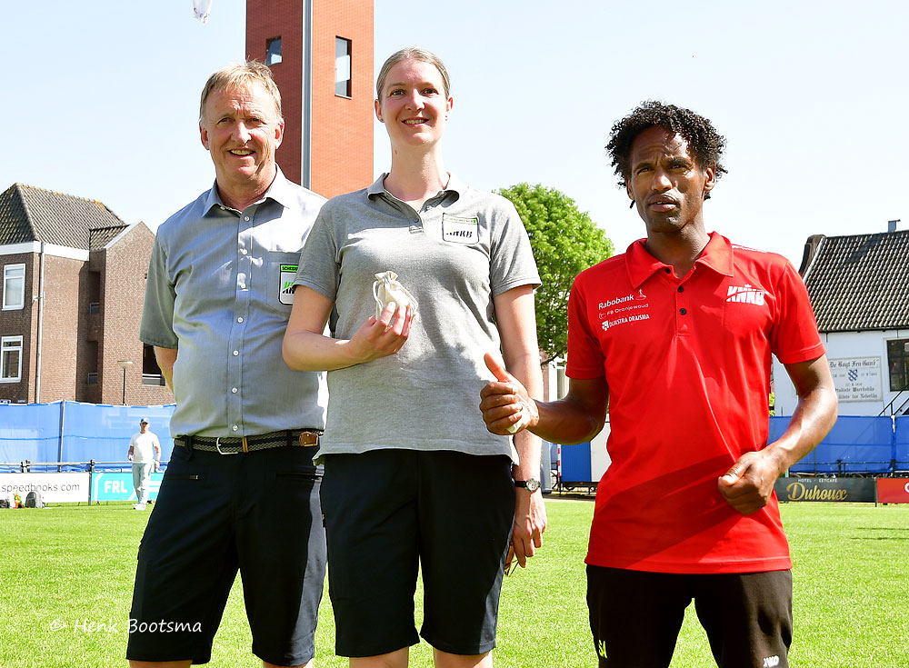 Three people stand on a sunny sports field posing for a photo: a man in a light blue shirt, a woman in a gray polo, and a man in a red polo.