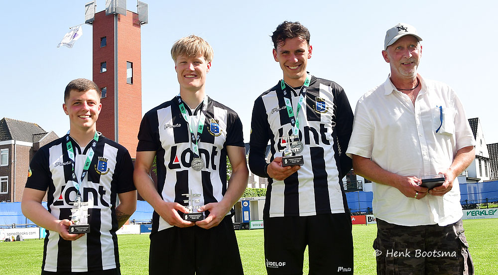 Four men on a sunny football field pose with medals and trophies; three players wear black-and-white striped jerseys, one man in a white shirt and cap stands at right.