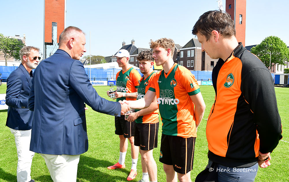 Official in a blue blazer shakes hands with a line of orange-and-green soccer players on a sunny field, likely an awards moment.