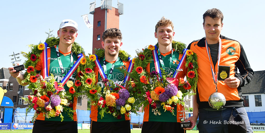 Four male athletes pose with colorful flower wreaths and medals after a victory, outdoors on a sports field.