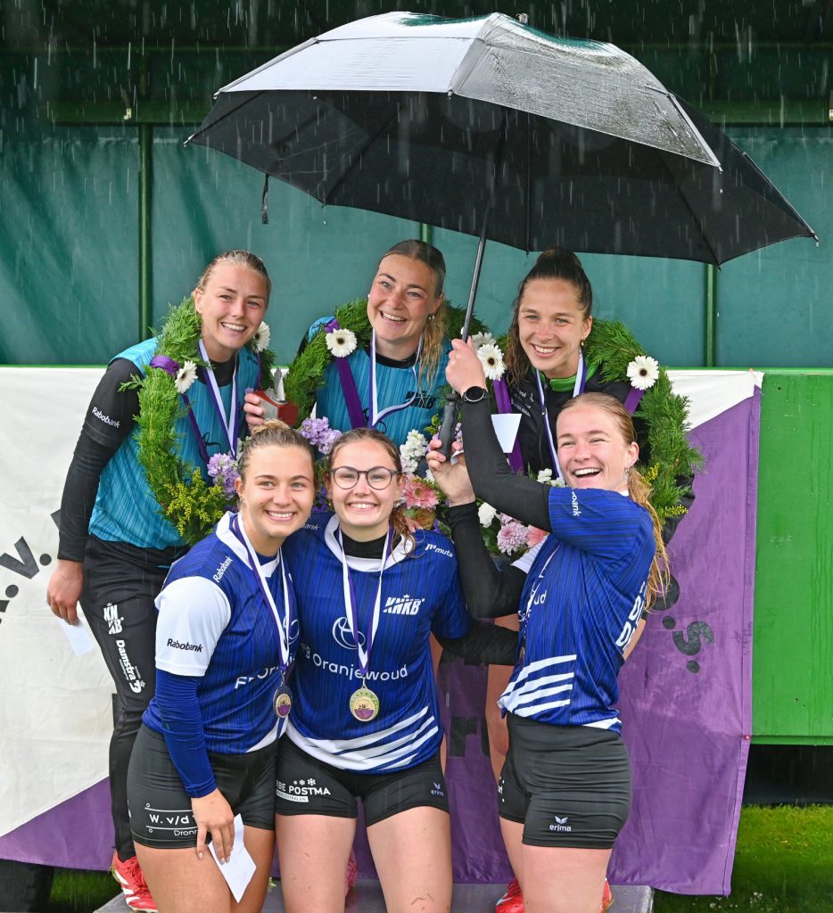 Dames Hoofdklas Totaal - kaatsnieuws.com Group of female athletes on a podium smiling, wearing blue jerseys with medals, holding a wreath and flowers in the rain under a large umbrella.