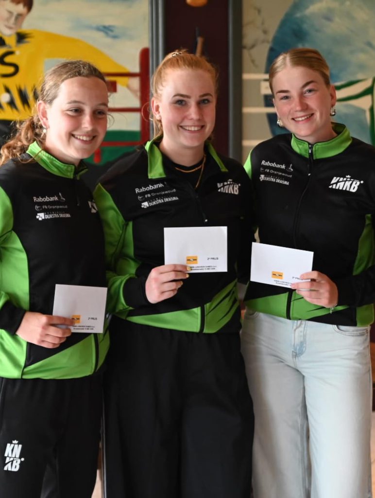 Dbroek2 - kaatsnieuws.com Three smiling women in black-green jackets holding white envelopes, posing for a group photo at an event