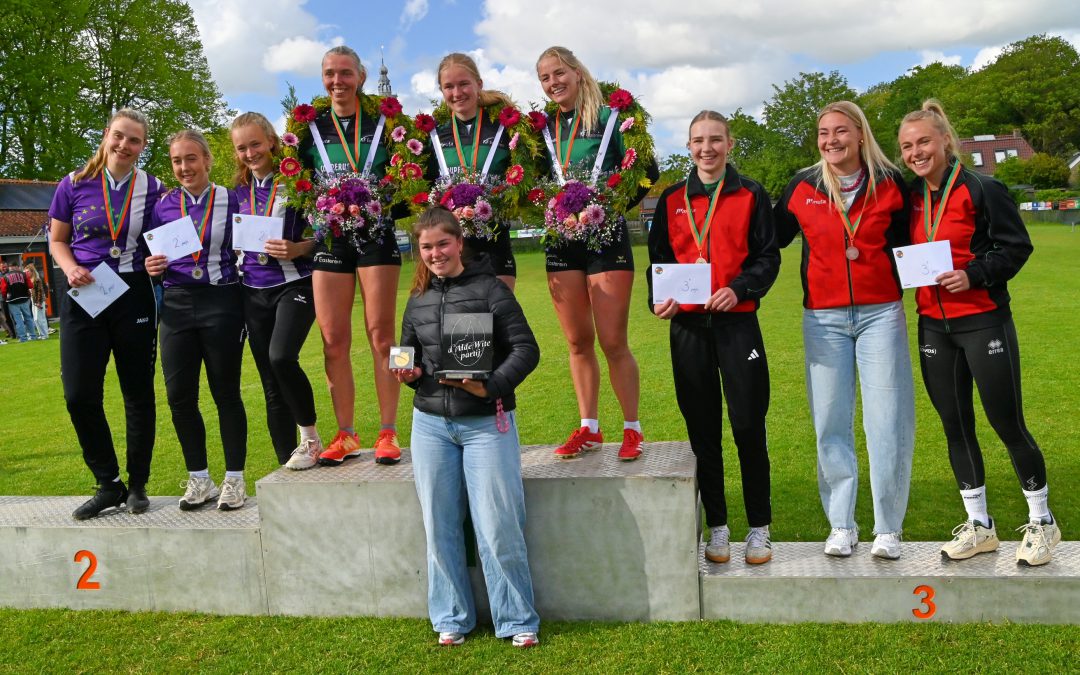 Group of smiling female athletes standing on a podium with medals and bouquets, posing for a photo outdoors on a sunny day.