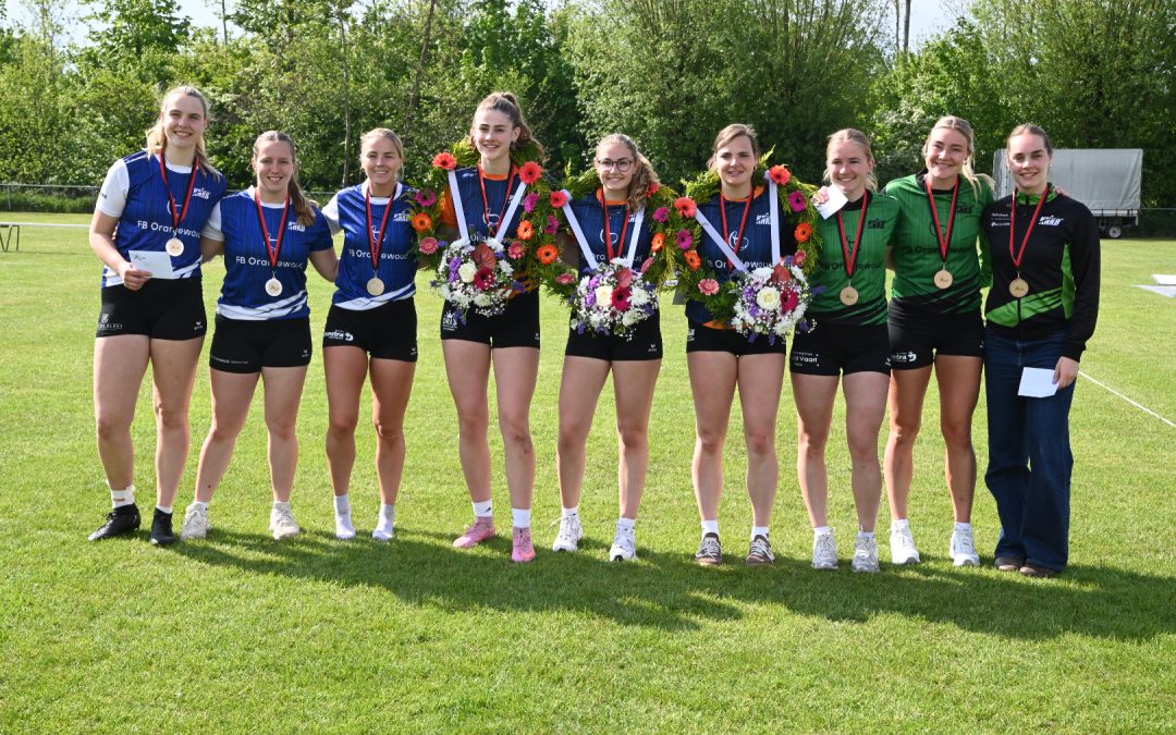 Nine young female athletes stand in a line on a sunny grassy field, wearing blue and green uniforms with medals and several holding colorful flower bouquets.