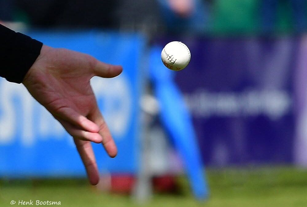 Close-up of a hand throwing a baseball; the ball is in midair with a blurred sports field in the background.