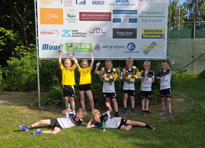 Young kids in yellow and black and white uniforms pose with trophies and flowers in front of a sponsor banner on a grassy field, celebrating a win.