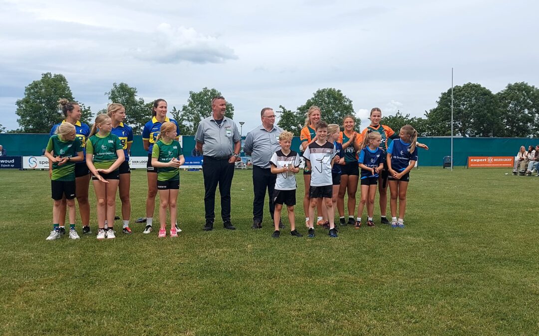 Group of young athletes in green, blue, and orange jerseys posing with two adult coaches on a grass field during an outdoor event.