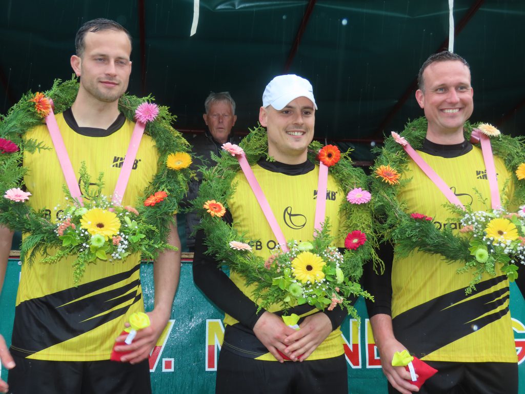 Minnertga heren 2e klasse vf Rutger Torensdm Riemer Hoerkstra en Hendrik Jan van der Velde - kaatsnieuws.com Three men in yellow and black outfits wearing flower garlands pose together, holding small items after a race ceremony.