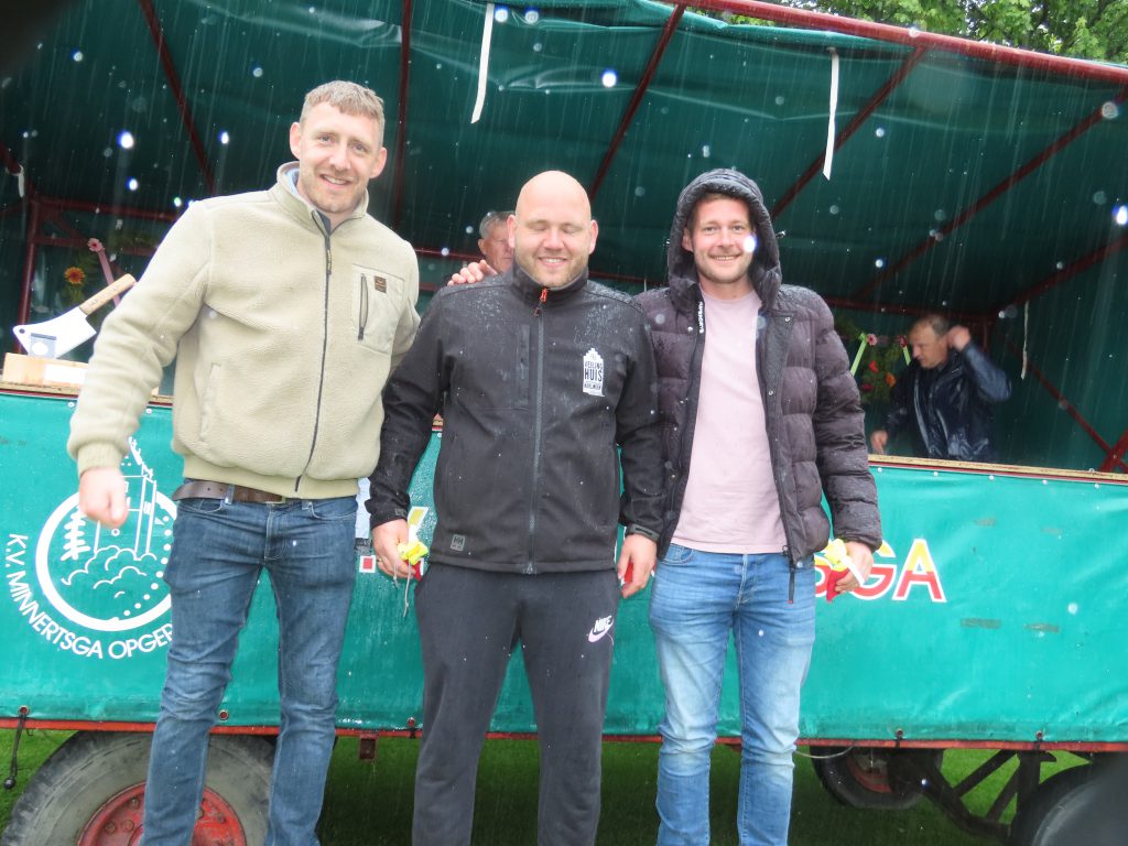 Minnertsga 17 mei 2026 heren 2e klasse vf derde prijs - kaatsnieuws.com Three men stand side by side outdoors, smiling in light rain in front of a green stall backdrop.