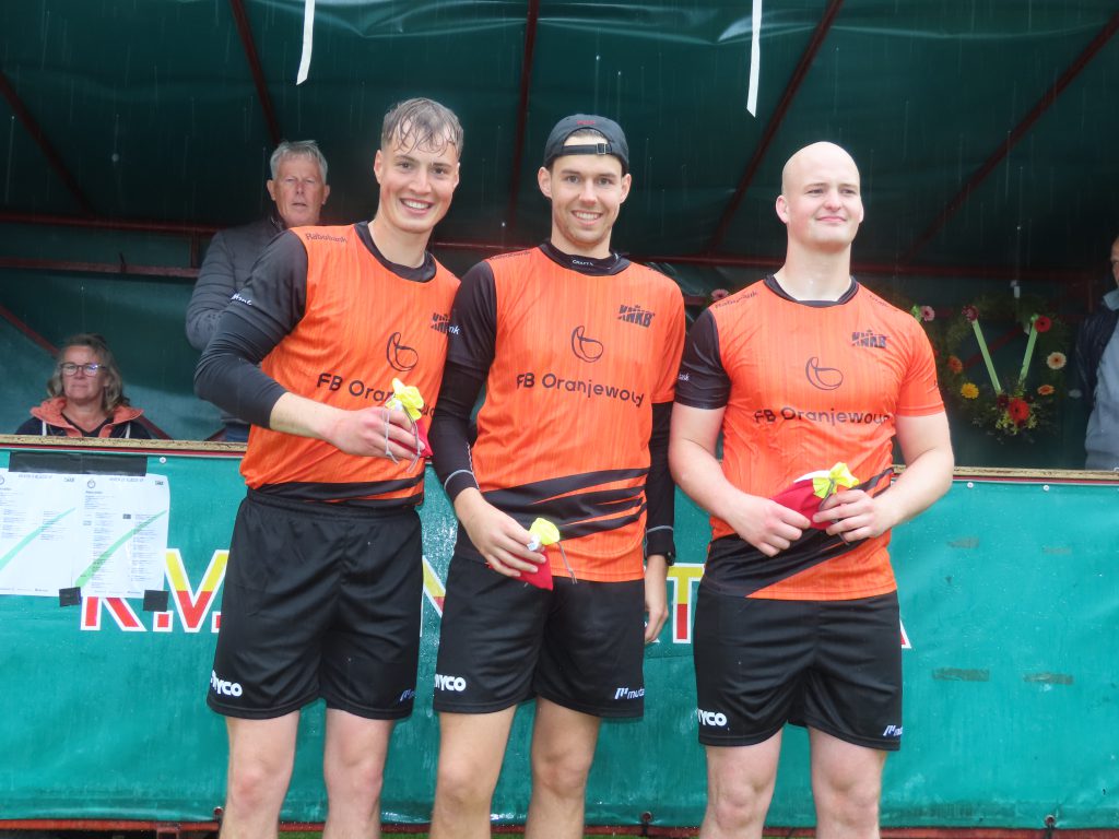 Minnertsga heren 1e klasse vf 17 mei 2026 Rick Minnesdma Jari Visser en Youri de Groot - kaatsnieuws.com Three male athletes in orange team jerseys standing on a podium, smiling and holding yellow bouquets, as light rain falls against a green backdrop.
