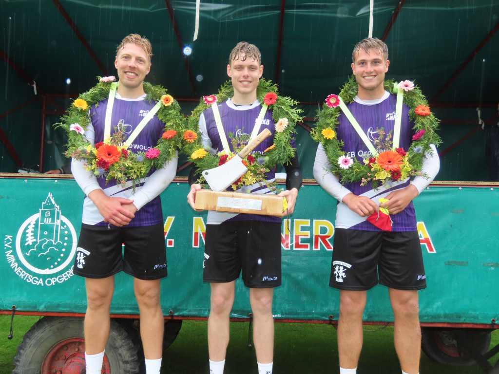 Minnertsga heren 1e klasse vf 17 mei 2026 Sjoerd de Jong Jorrit Nanninga koning en Jan Bandstra - kaatsnieuws.com Three male athletes in purple uniforms wear flower garlands and pose with a prize on a rainy outdoor stage. Behind them is a green backdrop.